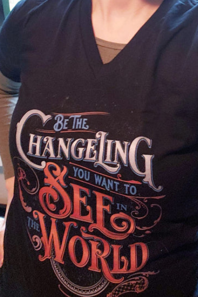A person wearing a black V-neck graphic t-shirt with the text 'Be the CHANGELING YOU WANT TO SEE IN THE WORLD' in white and red lettering.