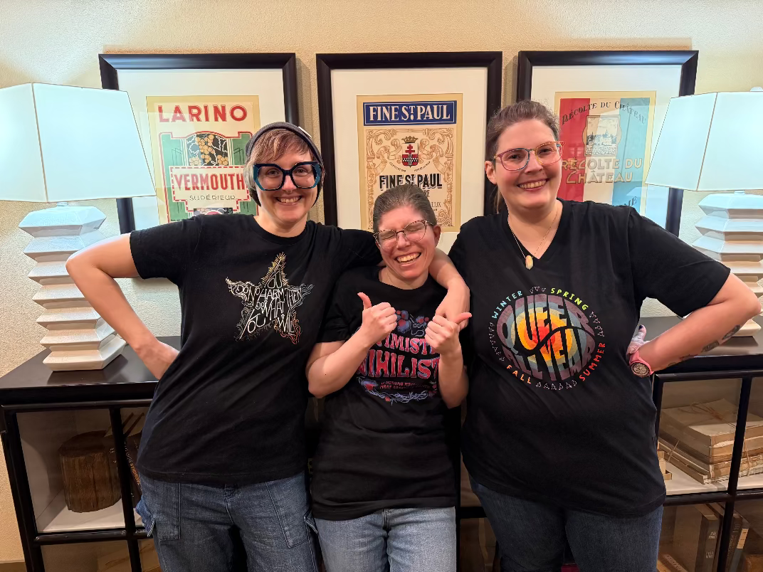 Three people wearing black t-shirts with colorful designs in a room with framed posters on the wall.