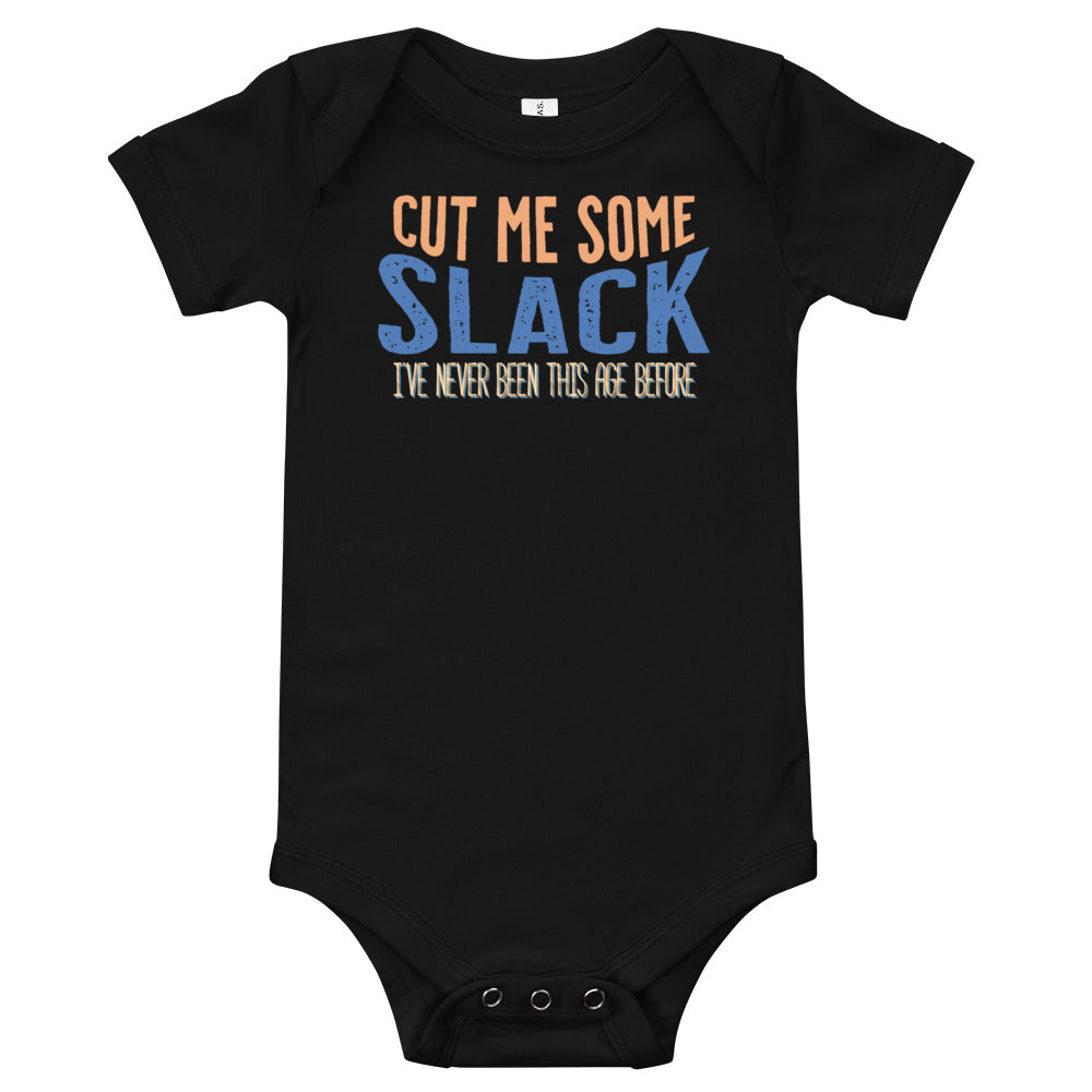 Black baby onsie with “Cut me some” in orange “Slack” in blue. “I’ve never been this age before” in smaller text underneath.