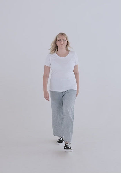 Video of white woman with blond hair showing the fit of the Relaxed Fem Fit Tee. It hugs curves, has a high crew neck neckline, sleeves can be a bit small on this, hemline flows below pants line.