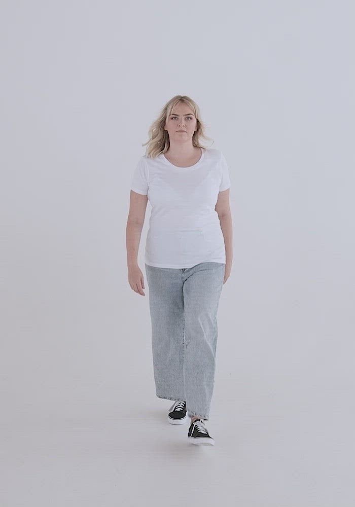 Video of white woman of medium build with blond hair showing the fit of the Relaxed Fem Fit Tee. It hugs curves, has a high crew neck neckline, sleeves can be a bit small on this, hemline flows below pants line.