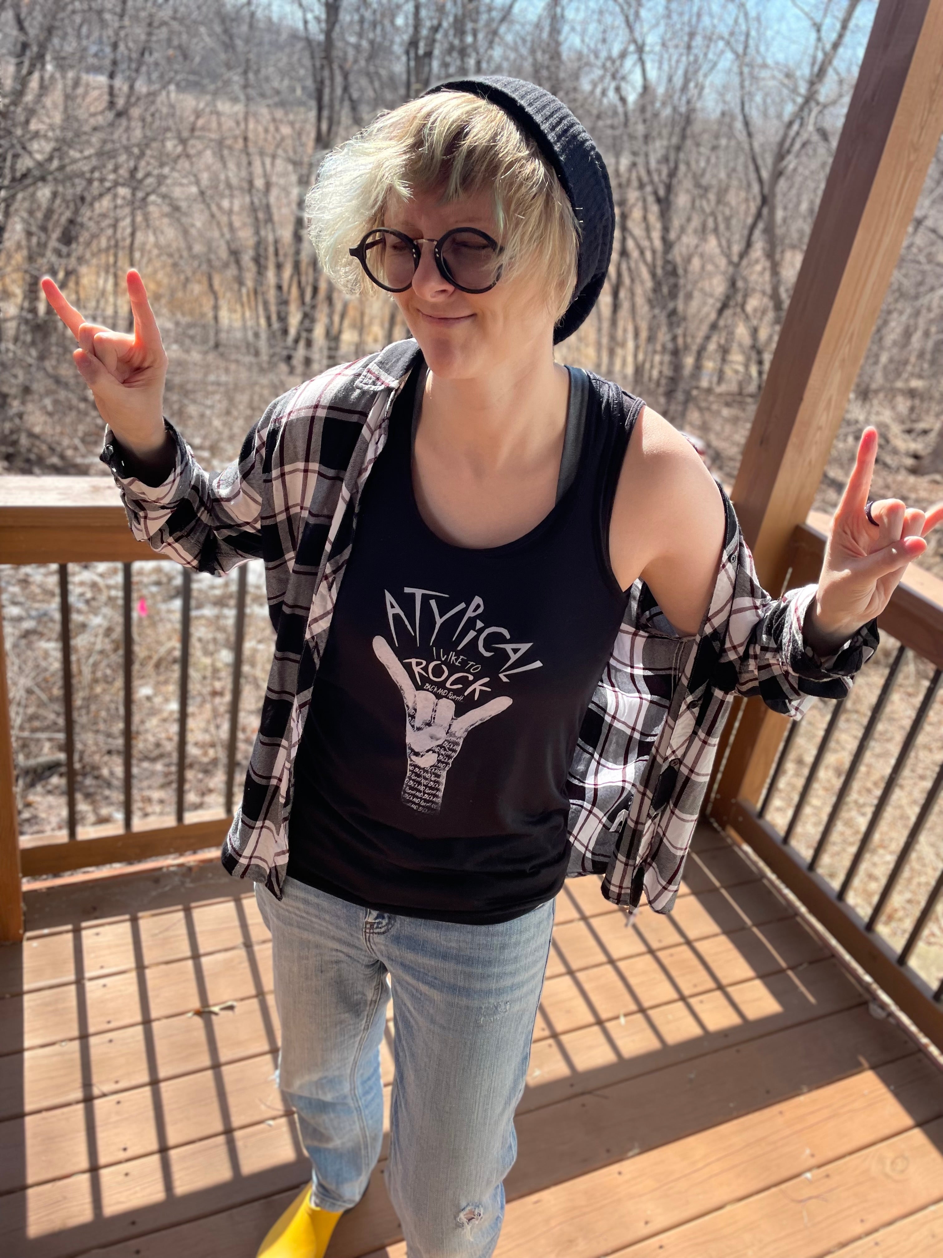 Lifestyle image of a white non binary model with slim build, glasses, slouch beanie hat wearing the black flowy racerback tank with design “Atypical, I like to ROCK”. Hands are both up in the “ROCK” gesture. 