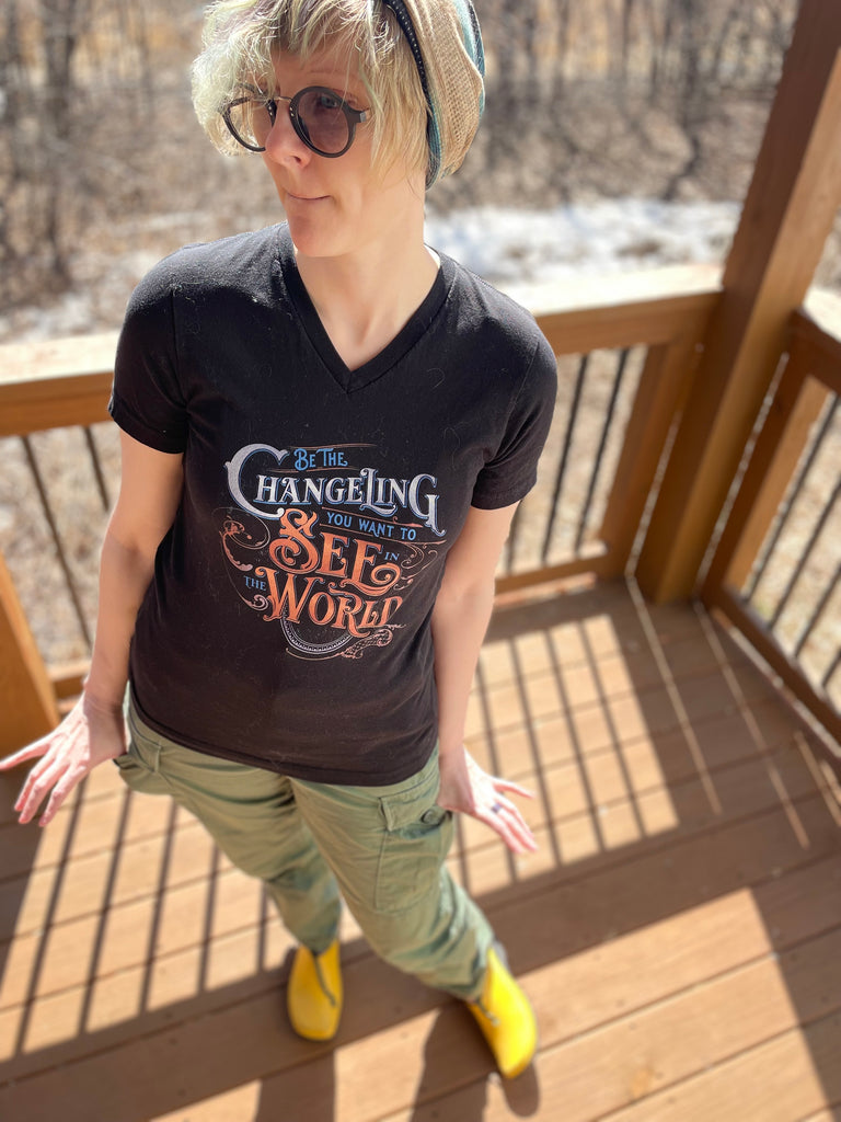 Lifestyle image of non binary model outside wearing glasses, a beanie hat and a black V-Neck unisex shirt with design  “Be the Changeling you want to see in the world” in silver, bronze and blue lettering surrounded by intricate decals