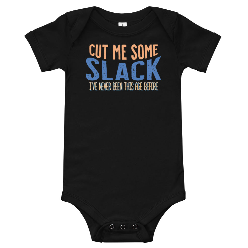Black baby onsie with “Cut me some” in orange “Slack” in blue. “I’ve never been this age before” in smaller text underneath. 