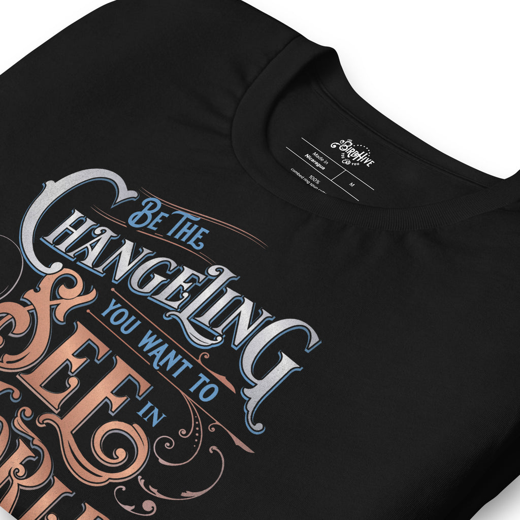 folded Black unisex fit tee with design  “Be the Changeling you want to see in the world” in silver, bronze and blue lettering surrounded by intricate decals